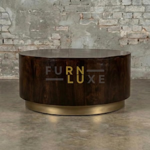 May include: A dark brown, cylindrical coffee table with a gold-coloured base. The table has a flat top and the text "FURN LUXE" is visible on the side. The table is set against a brick wall.