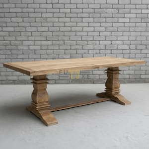 Op de afbeelding: Een rechthoekige eettafel van lichtbruin hout. De tafel heeft een massief blad en wordt ondersteund door twee sierlijke voetstukken, verbonden door een balk. De tafel staat tegen een grijze bakstenen muur. De tafel is ongeveer 200 cm lang.