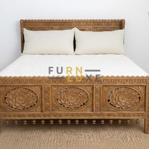 Puede incluir: Un marco de cama de madera con intrincadas tallas florales. El cabecero y el pie de cama presentan diseños detallados, y la cama está hecha de madera de color marrón claro. Dos almohadas blancas descansan sobre la cama, cubierta con una sábana blanca.