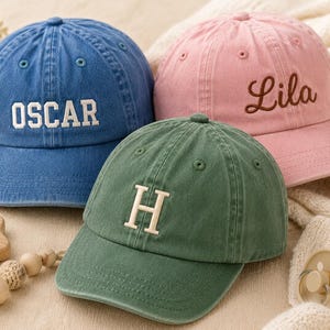 May include: Three baseball caps in blue, pink, and green. The blue cap features the white embroidered text OSCAR, the pink cap has the brown embroidered name Lila, and the green cap displays a large white embroidered letter H on the front.