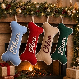 May include: Four bone-shaped Christmas stockings in light blue, red, beige, and dark green. Each stocking features white or dark red embroidered script names: Ethan, Ava, Chloe, and Liam. The stockings have white piping edges and hanging loops.
