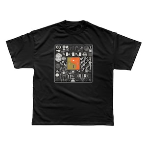 Puede incluir: Camiseta negra con un diseño gráfico cuadrado que contiene varios símbolos y un diseño central de un cuadrado naranja con un símbolo yin-yang amarillo y verde. La camiseta es de cuello redondo.