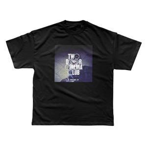 以下が含まれることがあります： 黒いTシャツ。正方形のグラフィックには、白い文字で「TWO DOOR CINEMA CLUB」と円形のデザインが描かれています。テキストの下には「FOREST HILLS, NY」と書かれています。このTシャツはクルーネックです。