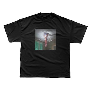 以下が含まれることがあります： 天井を突き抜けて落ちる人物のグラフィックが描かれた黒いTシャツ。「TWO DOOR CINEMA CLUB」と「Beacon」の文字が入った正方形の画像。Tシャツはクルーネックです。