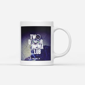 以下が含まれることがあります： 白いセラミックマグカップ。白で「TWO DOOR CINEMA CLUB」の文字、猫の目のグラフィック、下に「TOURIST HISTORY」の文字。マグカップには湾曲したハンドルが付いています。