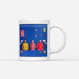 以下が含まれることがあります： 白いセラミックマグカップ。青色で「TWO DOOR CINEMA CLUB」の文字がプリントされています。デザインは、カラフルなトップスを着た3人の人物を特徴とし、メガホンや電話などのグラフィック要素を備えた青色の背景が特徴です。