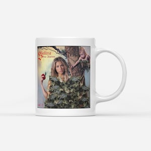 May include: White ceramic mug with a design featuring a woman holding an apple, and a baby in a tree. The mug has the text "Shakira Oral Fixation Vol. 2" in red and gold. The design is a stylized illustration.