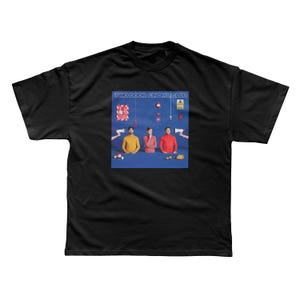 以下が含まれることがあります： Two Door Cinema Clubのアルバムアートワークがプリントされた黒いTシャツ。黄色、ピンク、赤のシャツを着た3人が青い背景に描かれています。バンド名は上部に印刷されています。