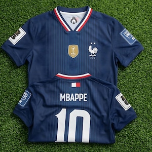 May include: A dark blue soccer jersey featuring a V-neck with red and white trim. The front displays a gold FIFA emblem and a white rooster logo with two stars. The back shows the name MBAPPE and the number 10 in white, with a small French flag.
