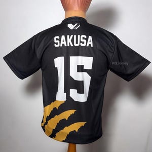 May include: Black jersey with the name "SAKUSA" and the number "15" in white. The jersey has a gold claw design on the lower left side and the text "KQ Jersey" on the right sleeve. A logo is visible at the top.
