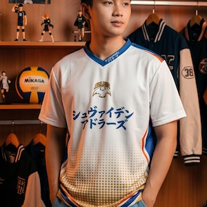 May include: A white V-neck jersey with blue and orange accents. The chest features the text "ADLERS" above a bird emblem and the Japanese text "シュヴァイデン アドラーズ". The lower portion has a gold-toned dot pattern, and the collar includes small Japanese text.