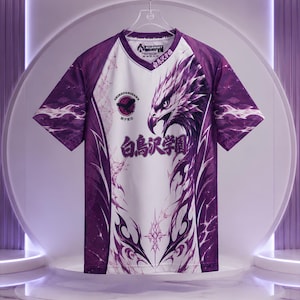 Puede incluir: Una camiseta de manga corta morada y blanca con un gráfico de cabeza de águila, patrones tribales intrincados y texto en japonés. La prenda incluye un logotipo circular con la silueta de un águila y el texto Shiratorizawa en el pecho.