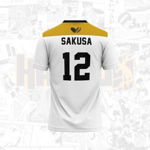 May include: White athletic jersey with a gold yoke and black accents. The jersey features the name "SAKUSA" and the number "12" in bold black lettering. A small logo is visible on the gold yoke.