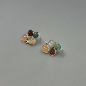 Könnte beinhalten: Ein Paar Ohrstecker mit einer Ansammlung farbiger Edelsteine. Jeder Ohrring hat eine silberne Basis mit goldenen Akzenten und eine Ansammlung runder Steine in Pfirsich-, Grün- und Brauntönen. Die Ohrringe sind auf grauem Hintergrund.