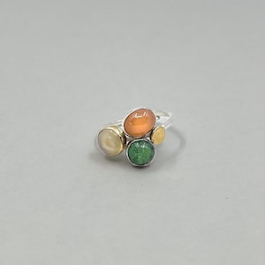 Puede incluir: Un anillo de plata con una disposición de piedras preciosas de colores. El anillo presenta un grupo de piedras naranjas, verdes, amarillas y blanquecinas. Las piedras están engastadas en una banda de plata, creando un diseño único y artístico.