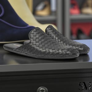 May include: A pair of black woven leather slip-on shoes. The shoes have a textured, basket-weave design and a rounded toe. The shoes are displayed on a dark surface, with other shoes visible in the background.