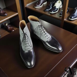 Puede incluir: Botas de cuero de dos tonos con la parte superior de tela gris y la parte inferior de cuero negro. Las botas tienen un diseño con cordones y perforaciones decorativas en la puntera. Se ven otros zapatos en los estantes del fondo.