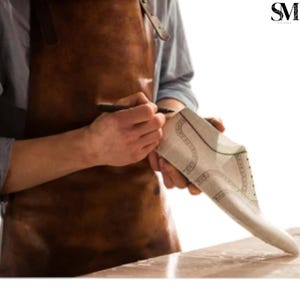 May include: A person wearing a brown leather apron is drawing on a shoe pattern with a black pen. The pattern is light-coloured with dark lines, showing the design of a shoe. The image has a warm, natural light.