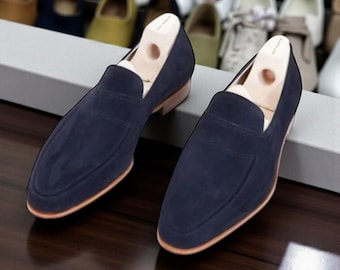 Può includere: Un paio di mocassini in camoscio blu navy con suole marrone chiaro. Le scarpe hanno un classico design slip-on e sono esposte con forme di legno. Altre scarpe sono visibili sullo sfondo.