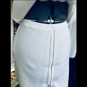 Op de afbeelding: Close-up van een witte rok met een lange rits. De rok is gemaakt van een textuurstof en wordt gecombineerd met een bijpassende witte top. De taille is zichtbaar, met het woord "WAIST" gedrukt op een zwarte band.