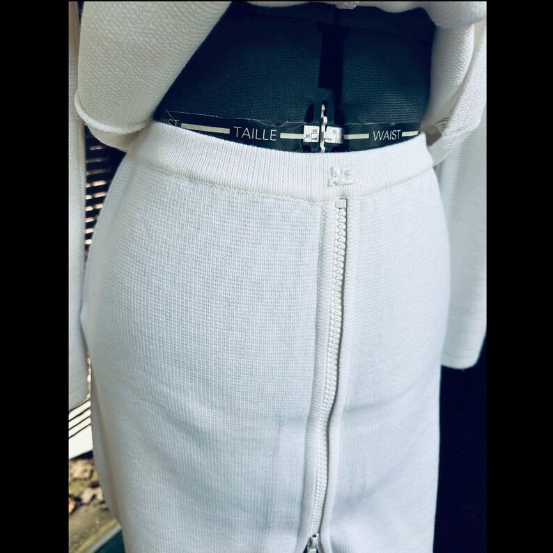 Op de afbeelding: Close-up van een witte rok met een lange rits. De rok is gemaakt van een textuurstof en wordt gecombineerd met een bijpassende witte top. De taille is zichtbaar, met het woord "WAIST" gedrukt op een zwarte band.
