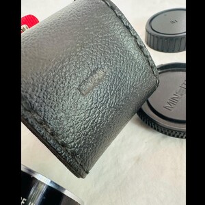 May include: A black leather camera case with a textured surface. Two black lens caps are visible, one with the brand name "MINOLTA" printed on it. The case is cylindrical and appears to be well-worn.