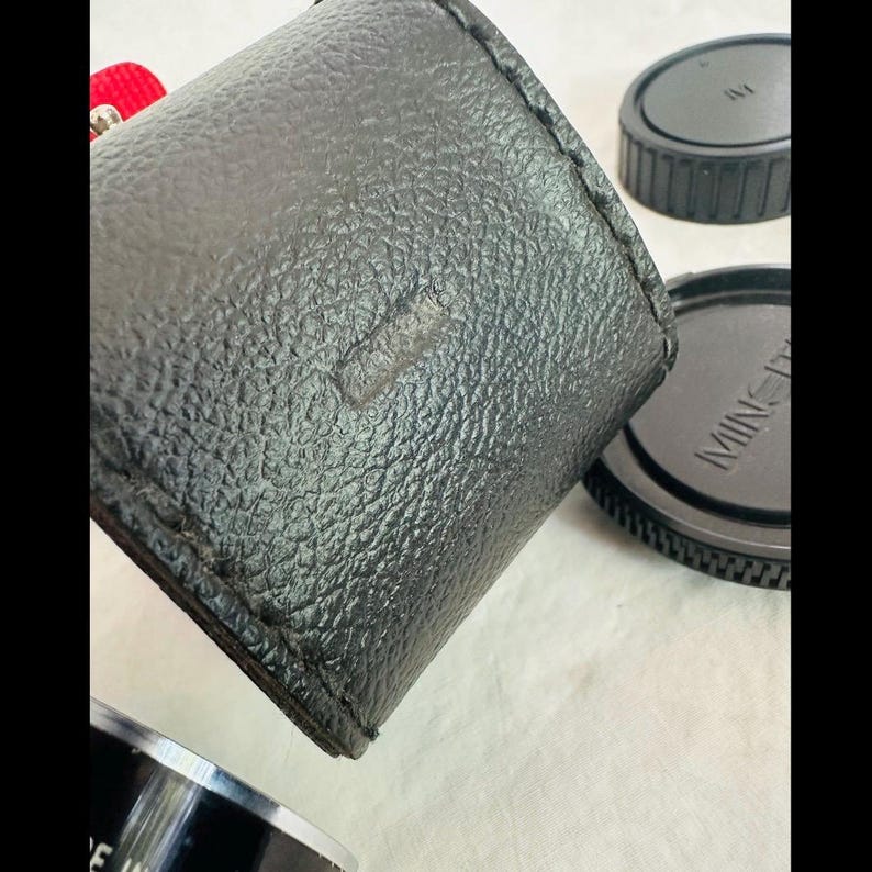 May include: A black leather camera case with a textured surface. Two black lens caps are visible, one with the brand name "MINOLTA" printed on it. The case is cylindrical and appears to be well-worn.