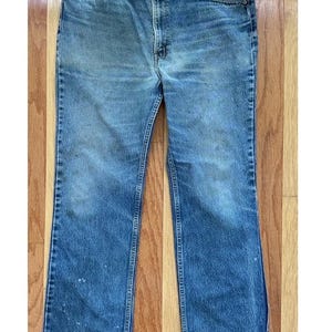 Könnte beinhalten: Eine mittelblaue Jeans aus Denim mit klassischem Fünf-Taschen-Design, Knopfleiste und geradem Bein. Der Stoff weist natürliche Verblassungen und leichte Abnutzungserscheinungen auf den vorderen Bereichen auf.