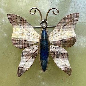 Puede incluir: Broche de mariposa dorada con intrincados detalles en las alas. El cuerpo de la mariposa presenta una piedra ovalada de color azul oscuro. El broche tiene un cierre de alfiler en la parte posterior, adecuado para asegurar a la ropa o accesorios.