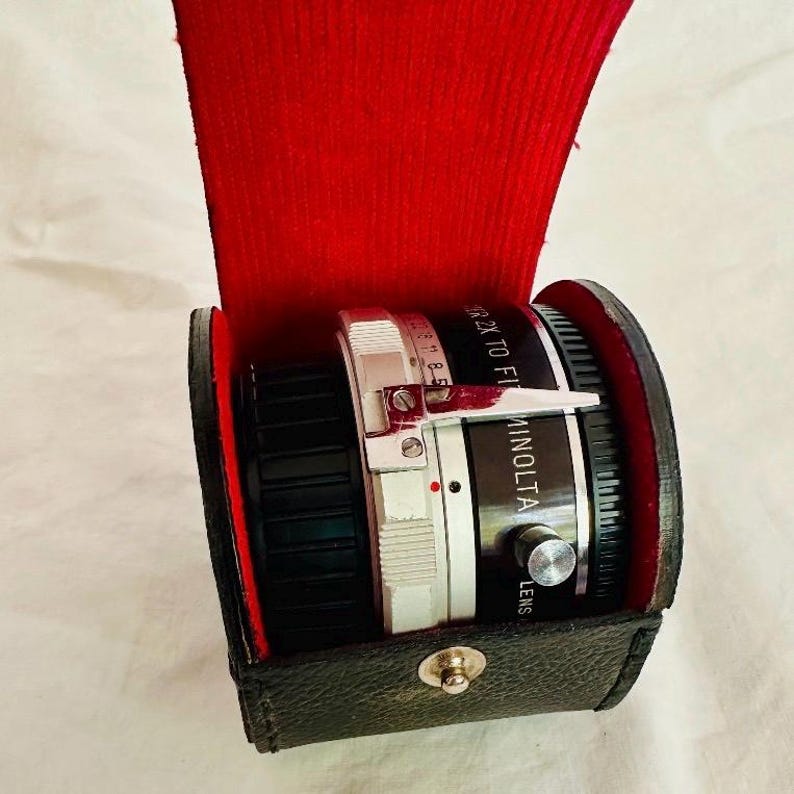 May include: A Minolta 2x teleconverter lens in a black leather case with a red velvet interior. The lens is silver and black with white text. The case has a silver clasp.