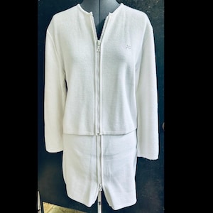 Op de afbeelding: Een tweedelige witte gebreide outfit met een lange ritssluiting op de jas en rok. De jas heeft lange mouwen en een klein logo op de borst. De rok is knielang. De outfit wordt op een mannequin getoond.