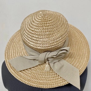 Puede incluir: Un sombrero de paja de ala ancha con un gran lazo de tela beige. El sombrero es de color beige claro y tiene una textura tejida.