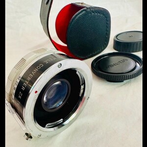 May include: A silver Minolta Tele Converter 2X lens with black lens caps and a black leather case with a red interior. The lens has a series of numbers and text around the edge.