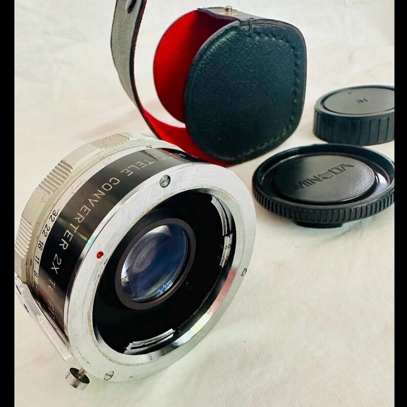 May include: A silver Minolta Tele Converter 2X lens with black lens caps and a black leather case with a red interior. The lens has a series of numbers and text around the edge.
