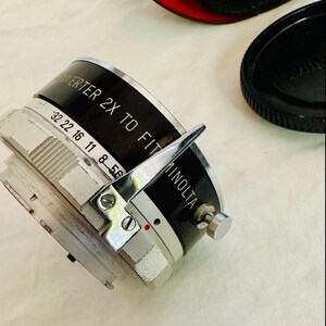 May include: A Minolta 2X to Fit camera lens converter with black and silver detailing. The converter features a silver adjustment lever and numbered markings. The word "MINOLTA" is printed on the side.