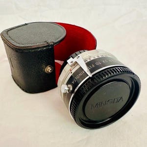 May include: A vintage Minolta 2x teleconverter lens in black and silver, alongside its black leather case with a red lining. The lens features a silver adjustment lever, and the Minolta logo is displayed on the lens cap.