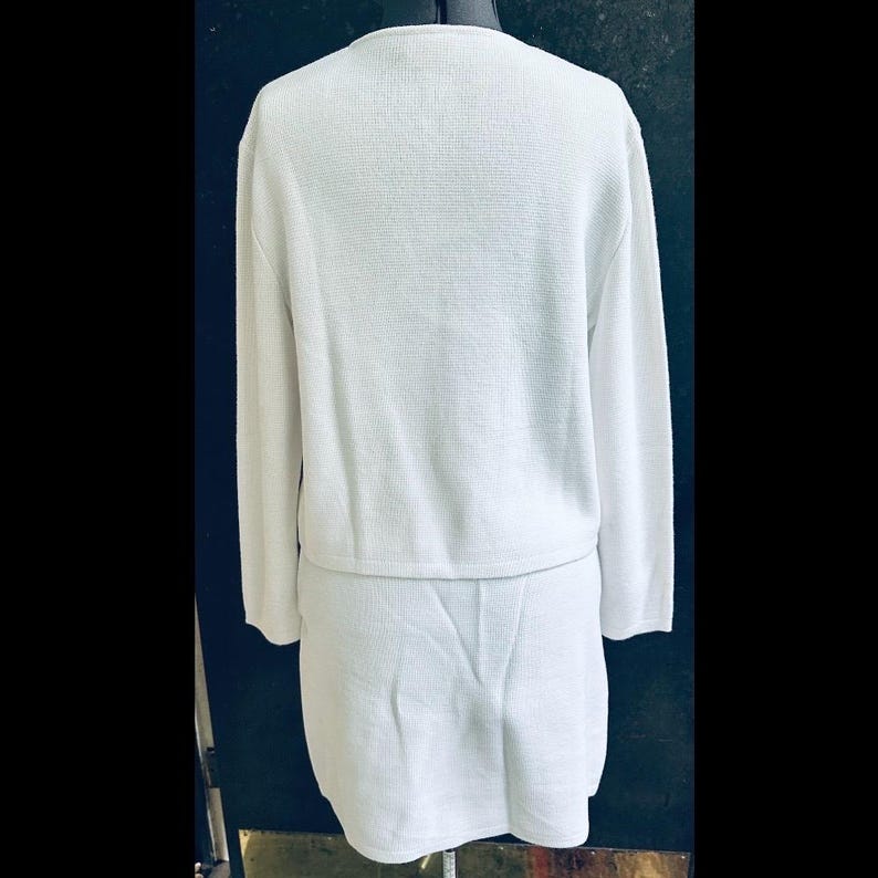 Op de afbeelding: Een tweedelige witte gebreide outfit, bestaande uit een top met lange mouwen en een bijpassende rok. De top heeft een ronde hals en de rok is knielang. De stof heeft een getextureerd uiterlijk. De outfit wordt op een mannequin getoond.