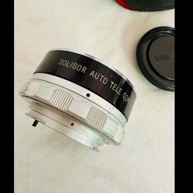May include: A Soligor Auto Tele Converter lens with a black and silver finish. The lens has the text "SOLIGOR AUTO TELE CONV" printed on the black band. A black lens cap with the text "MINOLTA" is also visible.