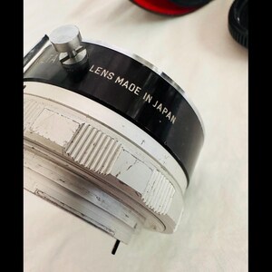 May include: A close-up of a camera lens with the text "LENS MADE IN JAPAN" printed on the black band. The lens has a silver and black colour scheme with a textured, ridged design.