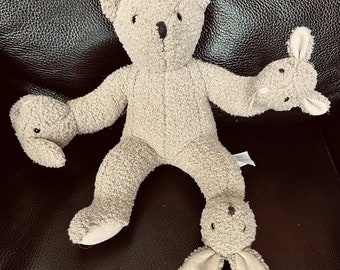 May include: A beige teddy bear with two beige bunny rabbit plush toys attached to its arms and legs. The teddy bear is sitting on a black leather surface.
