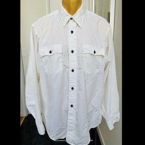 Può includere: Una camicia bianca a maniche lunghe con bottoni e due tasche sul petto con bottoni neri.