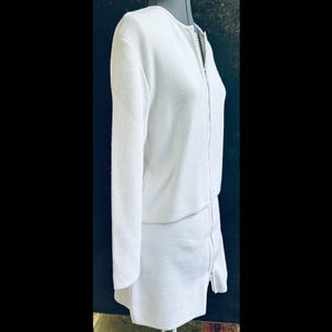 Op de afbeelding: Een witte gebreide tweedelige outfit, bestaande uit een jas met lange mouwen en een volledige ritssluiting en een bijpassende rok. De outfit wordt op een mannequin gepresenteerd tegen een donkere achtergrond. De stof heeft een textuur.