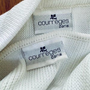 Op de afbeelding: Close-up van twee witte stoffen labels met de zwarte tekst "courr&egrave;ges paris". De labels zijn bevestigd op een witte gebreide stof. Het Courr&egrave;ges-logo staat boven de tekst.