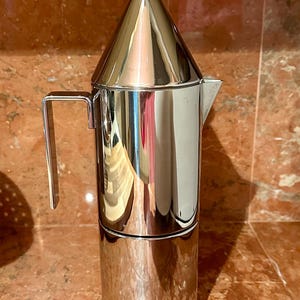 May include: A polished stainless steel stovetop espresso maker with a cylindrical body, a conical lid topped with a small spherical knob, and a rectangular handle. The coffee pot features a short, pointed spout and a reflective metallic finish.