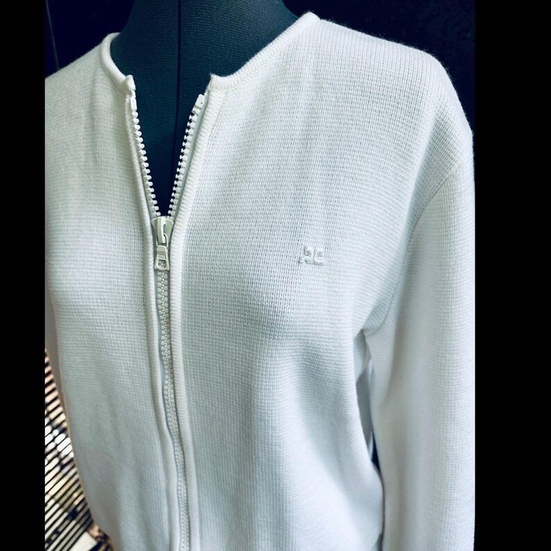 Op de afbeelding: Een witte trui met ritssluiting en een textuurgebreid patroon. De trui heeft een hoge kraag en een zilveren ritssluiting. Een klein logo is op de borst geborduurd. De trui wordt getoond op een zwarte mannequin.