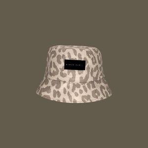 Peut inclure: Un chapeau bob beige avec un motif léopard. Le chapeau comporte un patch rectangulaire noir avec du texte blanc. Un accessoire de mode décontracté.