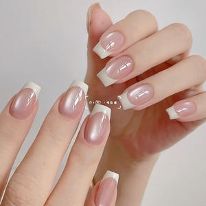 Può includere: Primo piano di una mano con manicure alla francese. Le unghie sono rosa pallido con una punta bianca. Le unghie sono lunghe e squadrate, con una finitura lucida. L'aspetto generale è elegante e classico.