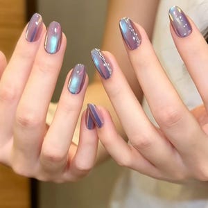 May include: Close-up of hands with oval-shaped, iridescent purple nail polish. The polish shifts colours, displaying hints of blue and pink. The nails are neatly manicured, reflecting light and creating a shimmering effect. The overall aesthetic is elegant and modern.
