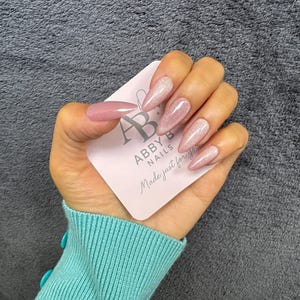Puede incluir: Una mano con uñas artificiales largas en forma de almendra. Las uñas son una mezcla de malva sólido y rosa brillante. En la mano se sostiene una tarjeta de visita con el texto "ABBY B NAILS Made just for you". La persona lleva un suéter turquesa.