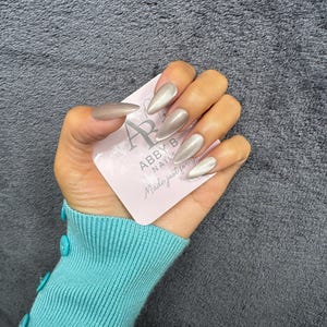 May include: A hand with long, almond-shaped, metallic taupe-colored artificial nails. The nails are glossy and reflect light. The hand holds a pink card with the text "ABBY B NAILS Made just for you."